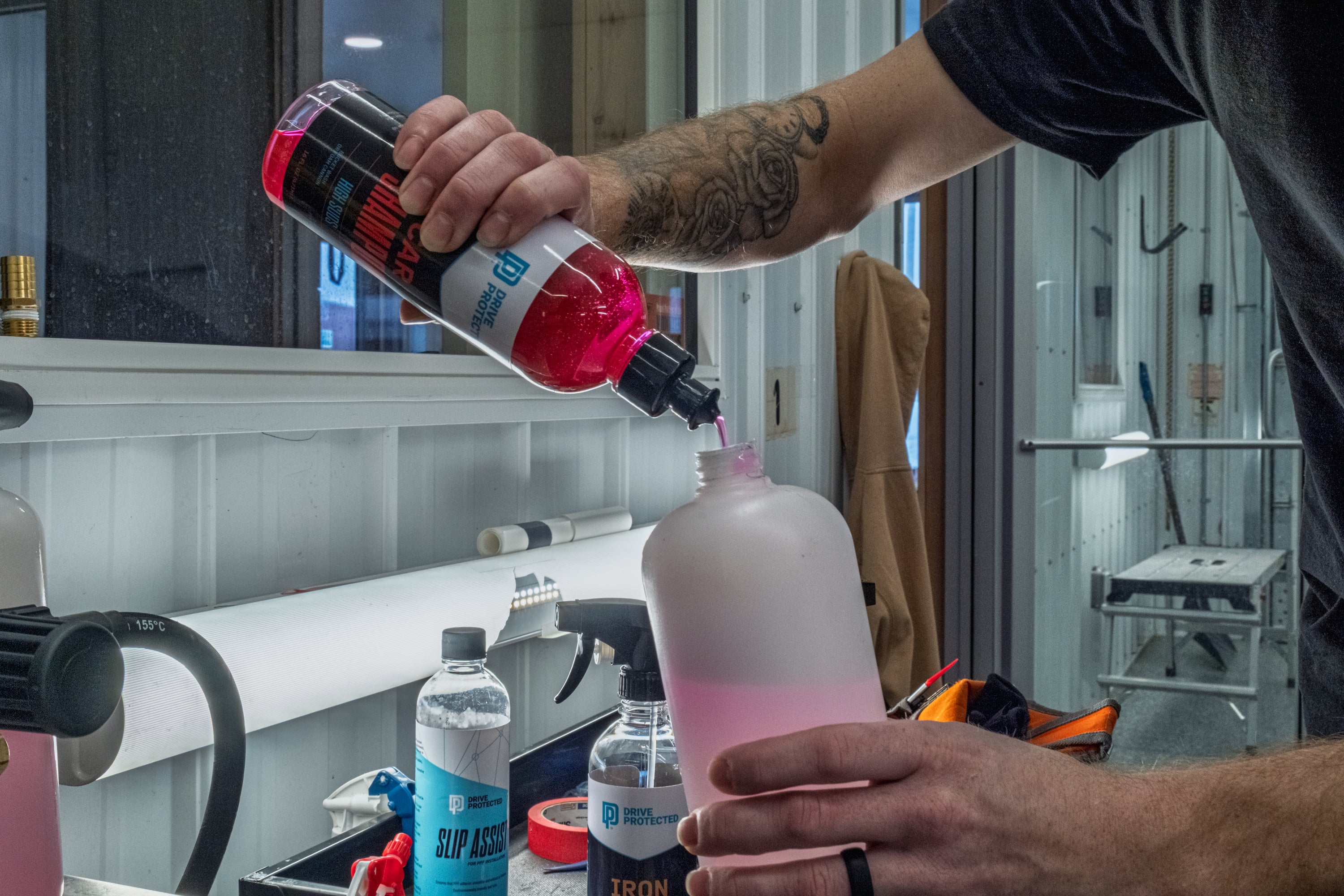 Person pouring liquid from a red and black bottle into a pink bottle with a white background.