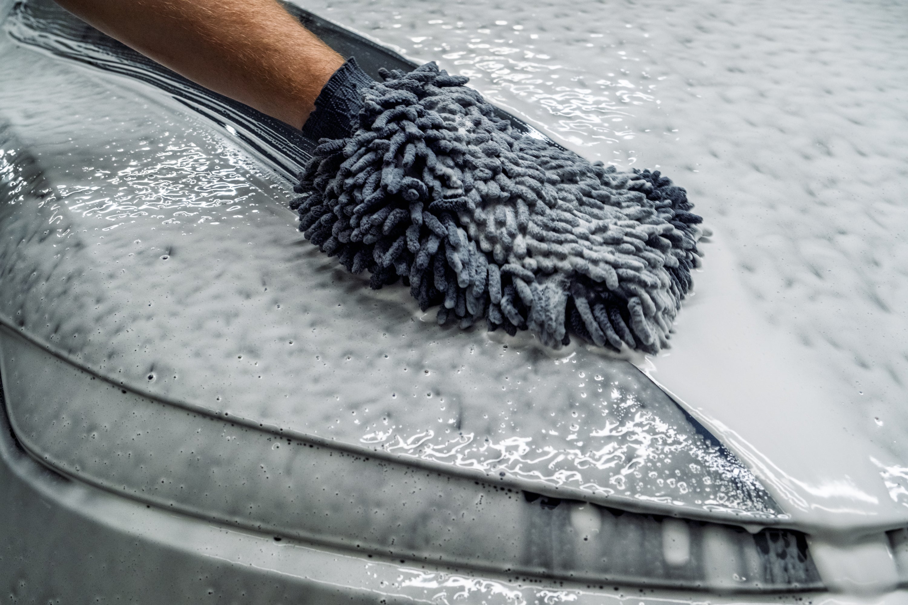 Person cleaning a car with a gray microfiber mitt on a soapy surface